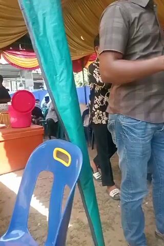The video shows an outdoor event, likely a gathering or celebration, taking place under a yellow and red tent. Several people are present, seated on blue plastic chairs and standing. The ground is covered with dirt and some grass. In the foreground, a man in a brown checkered shirt and blue jeans walks past a large cylindrical pole. Further into the scene, a young boy in a black shirt with a floral pattern drinks from a cup. Other individuals are visible in the background, some seated at tables, suggesting a meal or social interaction. The architecture of a wooden building is visible behind the tent, with windows suggesting daylight. The overall atmosphere appears to be casual and communal. No specific text is clearly legible. The location is identified as Blanggurah, Indonesia. The time of day appears to be daytime, with no clear indication of weather conditions.