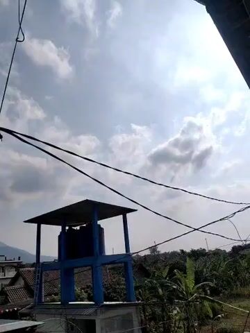 The video shows an outdoor scene with a clear blue sky and scattered clouds. In the foreground, there are several power lines running diagonally across the frame. To the left, a water tower with a blue tank and a dark roof stands on a concrete structure. In the background, there are rooftops of houses, lush green trees, and a mountain range in the distance. The sun is shining, casting light on the scene. The camera angle is slightly upwards, looking towards the sky and the objects in the environment.