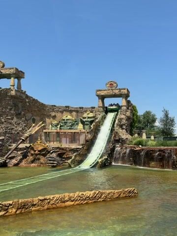 A group of people ride a water flume ride. The boat is going down a steep drop creating a large splash. The ride is themed to an ancient Egyptian temple. The weather is sunny and clear.