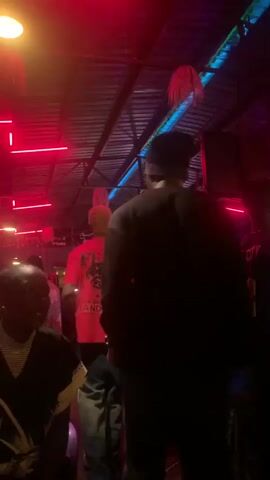 A dimly lit, indoor venue with red and blue neon lights illuminates a crowd of people. In the foreground, a person wearing a black shirt with a white design sits facing the right, their face partially obscured. Behind them, a person in a black long-sleeved shirt and a beanie walks away from the camera. Other individuals are visible in the background, some wearing red shirts or white and red striped tops. The ceiling appears to be made of corrugated metal, with decorative hanging elements. The overall atmosphere suggests a social gathering or entertainment venue.
