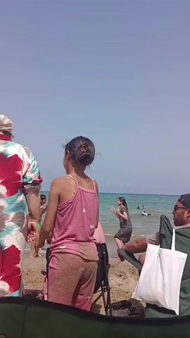 The video opens with a young girl in a pink tank top and patterned pants standing on a sandy beach, facing away from the camera. She is looking towards the ocean, where other people are visible. Behind her, a man in a green camping chair is looking off to the side. A white bag is resting on the sand in front of the man. Further back, a woman in a dark patterned dress is walking near the water's edge, holding a yellow object. A woman in a black full-body swimsuit stands near the ocean, looking down. Several beach umbrellas are visible, including a blue and white striped one in the foreground and a white one with scalloped edges behind it. Other beachgoers are scattered across the sand and in the water, some sitting under umbrellas and others swimming. The sky is clear and blue.