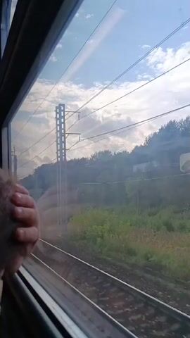 The video is shot from inside a moving train, looking out the window. A person's hand is visible resting on the window frame on the left side of the screen. Outside, train tracks stretch into the distance, flanked by green vegetation and trees on a gentle slope. Overhead, power lines and supporting structures for an electrified railway are visible against a partly cloudy sky. The sun appears to be casting a warm light, creating some atmospheric haze. The train is clearly in motion, as evidenced by the blurred scenery and the perspective shift.