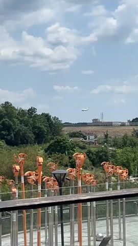 A light airplane flies high in a cloudy sky above a green landscape. In the foreground, a row of bright orange kinetic sculptures stand tall on poles, their fan-like elements catching the wind. A metal railing separates the viewer from the sculptures. An airport runway or landing strip is visible in the mid-ground.