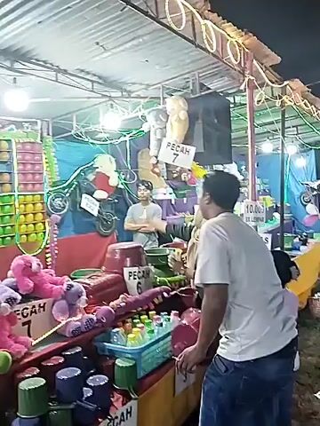 At night, a carnival game booth is shown. A man in a white t-shirt stands looking at the booth while two other customers approach from the side. The booth has a metal roof and is decorated with strings of light. The game consists of throwing balls to break balloons in a wall. Stuffed animals are displayed as prizes, including pink teddy bears and green caterpillars. There are also bottles of liquid. Two signs read "PECAH 6" and "PECAH 7". A man is standing behind the booth with his arms crossed.