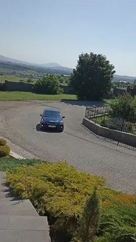 A blue car drives down a paved road. The road curves around a manicured garden with trimmed bushes and trees. In the background, a white church with a gold dome and cross is visible amongst the trees. The sky is clear and blue.