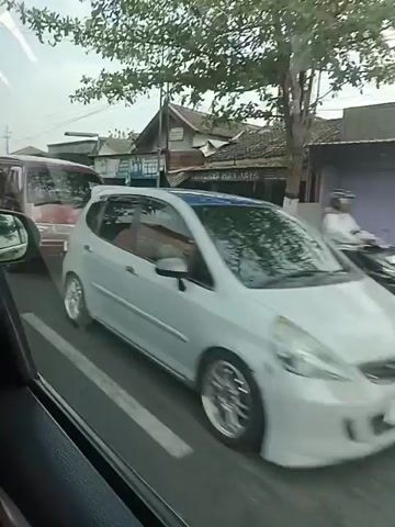 The video captures a series of vehicles moving along a road. The perspective appears to be from inside another vehicle, looking out the side window. The first vehicle visible is a maroon van. Following this is a silver hatchback, then a silver SUV. A yellow truck with a load of what appears to be wood or debris is next, followed by a white sedan. The environment seems to be a town or rural area with houses and shops lining the road. Power lines are visible overhead. The lighting suggests it is daytime, possibly overcast. The road is paved and appears to be a two-lane road. Other vehicles, including a motorcycle, are also briefly seen in the background or passing by. The movement of the vehicles indicates traffic is flowing.