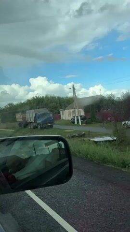 The video appears to be filmed from the passenger seat of a moving vehicle, looking out the side window and into the side mirror. In the side mirror, a person wearing a white shirt is visible. Outside the vehicle, a large blue truck with a trailer is parked on the side of a road. The truck is loaded with what appears to be agricultural produce or materials. To the right of the truck is a house with a red roof and beige walls. Trees and green vegetation line the road. The sky is blue with scattered white clouds. Several traffic signs are visible along the road. The overall impression is a rural setting.