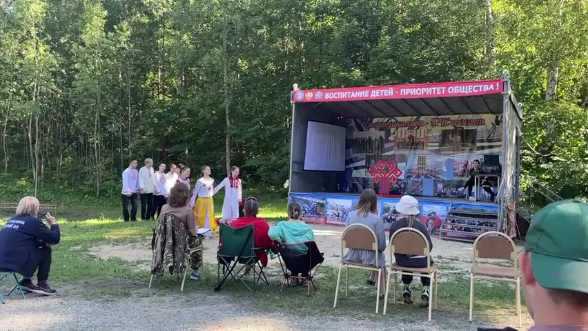 This video captures an outdoor performance or ceremony taking place in a wooded area. The scene is set on a makeshift stage, possibly a mobile stage unit, with a backdrop featuring colorful images and text. On the stage, a group of individuals, including children dressed in traditional clothing, are present. To the left of the stage, a line of young people in white shirts and dark pants stand at attention.

In front of the stage, an audience is seated in portable chairs, facing the performers. Some audience members are visible from behind, while one person in the foreground is wearing a green cap. A person in the lower left corner is holding a camera, appearing to record the event.

The weather appears to be clear and sunny, suggesting it's daytime, likely late morning or afternoon, given the lighting. The presence of trees indicates a natural, possibly rural or park-like setting. The overall atmosphere seems to be one of a community event, possibly a cultural performance or an educational program.

Visible text on the stage backdrop reads "ВОСПИТАНИЕ ДЕТЕЙ - ПРИОРИТЕТ ОБЩЕСТВА!" which translates to "CHILDREN'S EDUCATION - A PRIORITY FOR SOCIETY!". This text provides context for the event, suggesting it is related to child development and societal values. The imagery on the backdrop also appears to depict scenes of children, education, and possibly national or local symbols. The general mood seems calm and engaged, with the audience observing the proceedings attentively.