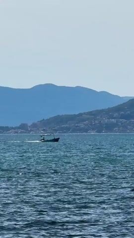 A small boat with a canopy is moving across a blue body of water. The boat has at least two people in it. In the background, there are hazy blue mountains with a city built on their slopes. The sky is a light blue. The water is choppy and reflects sunlight.