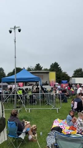 The video shows an outdoor event with a stage in the background. A crowd of people are seated on the grass in front of the stage, many of them in camping chairs or on blankets. Some people are standing and walking around. There is a blue tent in the foreground, and metal barriers. A surveillance camera is visible on a pole. The sky is overcast. In the middle ground, there are food stalls or vendor tents, and a bar area. Children are present, and one is sitting on a blanket with an adult. A dog is also visible on a blanket with a person. The overall scene suggests a festival or an outdoor concert.