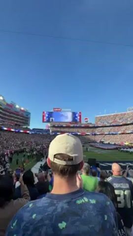 The video shows a view of a football stadium, packed with a crowd of spectators. The camera is positioned behind a spectator wearing a light-colored cap.  Overhead, a formation of military aircraft, including a larger central aircraft and smaller fighters, perform a flyover during the event. The video is shot on a bright, clear day with a blue sky.