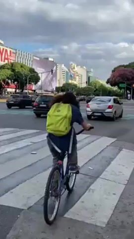 A person wearing a light-green backpack rides a bicycle across a crosswalk at an intersection in an urban setting. Cars are stopped at the intersection in both directions. The traffic light on the opposite side of the street is green. Buildings are visible in the background. Trees line the street, and one tree with pink blossoms is visible on the right.