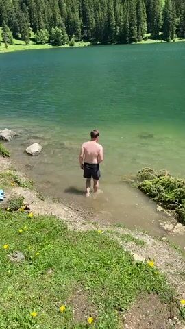 A man stands in the shallow, clear water of a lake, with his back to the camera. He is wearing black swim trunks. The lake is surrounded by a lush, green forest that climbs up a steep, rocky mountain. The sky is blue and clear. The water is a vibrant green color. There are some yellow flowers growing on the grassy bank in the foreground. The man appears to be about to enter the water further, or possibly just cooling his feet.