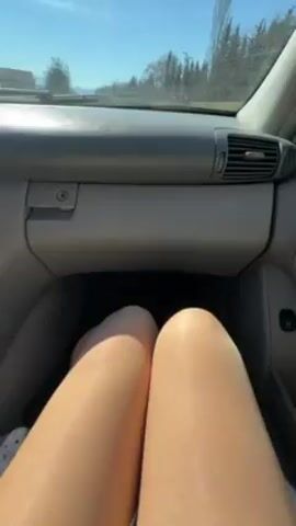 The video shows a car driving on a rural road on a sunny day. The camera is positioned inside the car, focused on the passenger side. Initially, the lower portion of a person's legs, wearing what appears to be pantyhose or stockings, is visible between the dashboard and seat. As the video progresses, the view shifts to show the road ahead through the windshield. There are trees and green fields on either side of the road. Another car is seen driving in the distance. The dashboard of the car is grey. The sky is bright blue with a few thin clouds.