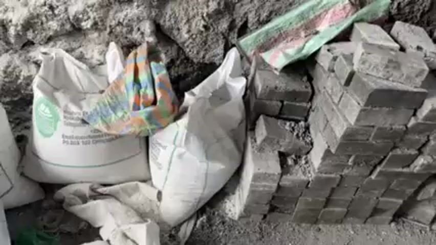 The video shows a pile of gray bricks stacked on the ground. Beside the bricks are two white bags and a multi-colored bag. The bags are lying on the ground and have some dirt on them. In the background is a gray, rocky wall. There is also a broom leaning against the brick pile.