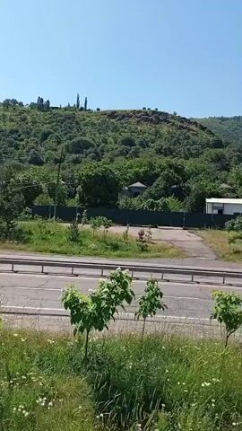 The camera faces a two-lane road with a metal guardrail separating it from the foreground, which consists of green grass and a few small trees. Behind the road, a densely wooded hillside rises, reaching a peak in the distance. Several buildings with light-colored roofs are nestled among the trees. The sky is clear and blue, indicating a sunny day.