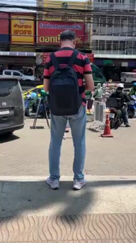 The video shows a man in a striped shirt, light blue jeans, and a dark backpack standing on a sidewalk, looking at his phone. He is in an urban environment with buildings and signs in the background. There are vehicles and other pedestrians passing by. The overall impression is that he is in a busy street or market area.