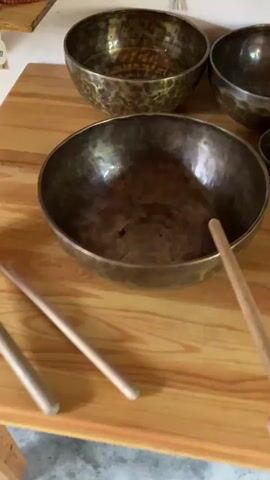 In this close-up, handheld video, a person's right hand is visible holding a wooden mallet. The mallet is about to strike a Tibetan singing bowl, which is positioned in the foreground of the frame. The bowl is made of a dark metal with a hammered texture and reflects light from its interior. Behind this main bowl, several other Tibetan singing bowls of varying sizes are arranged on a wooden surface, likely a table. These bowls are also dark and appear to have a similar hammered finish. The setting is indoor, with a plain wall visible in the background. The lighting suggests it's daytime, but no specific time or weather conditions can be determined. The activity is the preparation to strike a singing bowl, suggesting a moment of calm or anticipation, possibly for meditation or sound healing. There is no visible text in the video.