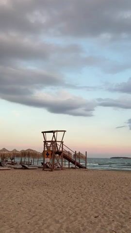The image presents a tranquil beach scene during what appears to be late afternoon or early evening, characterized by soft, diffused lighting. In the foreground, light brown sand stretches across the frame, showing some faint patterns.

A prominent wooden lifeguard tower stands centrally on the beach. It is a two-story structure with a viewing platform at the top, accessible by stairs on its right side. On the front of the tower, a blue square sign with a white cross is visible, commonly denoting a first aid or lifeguard station.

To the left of the lifeguard tower, several thatched beach umbrellas or cabanas are lined up, with sun loungers visible beneath some of them, indicating a commercial beach area.

Beyond the sandy shore, the ocean extends to the horizon, displaying gentle waves breaking near the coastline. A small, dark landmass or island can be seen in the distant right.

The sky above is a mix of light blue and soft grey clouds, with subtle hints of pink and orange near the horizon, typical of sunset or twilight. Despite the cloud cover, there are patches of clear sky. The overall atmosphere is quiet and serene, with no people visible in the frame, suggesting a calm moment on the beach. The setting is Voula, Greece.