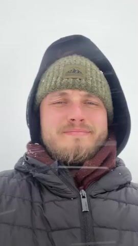 A man is taking a selfie outside in the snow. He is wearing a black puffer jacket, a green beanie, and a maroon scarf. A statue of a man holding a rifle stands in the background, covered in snow. The weather is snowy and overcast.