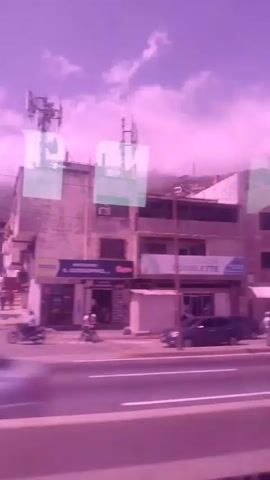 A video shows a view from inside a vehicle looking out at a street scene. Buildings line the street, including a multi-story brick building and a white building with a sign that reads "SOULETTE." Cars and a motorcycle are visible on the street and sidewalk. Several people are walking on the sidewalk. In the background, a hillside is covered with houses and vegetation. The sky is partly cloudy. The video has a pinkish hue.