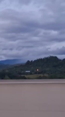 A slow pan across a cloudy sky over a lush green landscape. In the distance, a cluster of mountains stretches across the horizon. In the mid-ground, a dense forest covers rolling hills. A small, white building with a light emanating from it is visible in the center of the frame. A brown surface occupies the foreground, suggesting the perspective is from a vehicle or elevated structure.