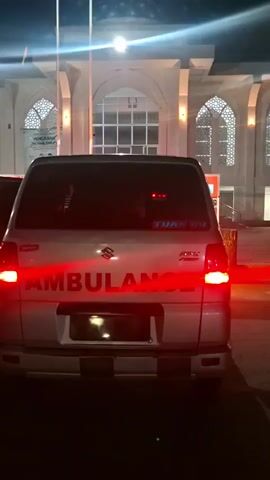 The video shows the rear of a white Suzuki ambulance parked in front of a large building with distinctive Islamic architectural features, including arched windows with intricate latticework. The building is illuminated by lights, suggesting it is nighttime or late evening. The ambulance has "AMBULANCE" written in large red letters across its back. A red light bar is active on the roof of the ambulance, emitting a bright red glow that illuminates the rear of the vehicle and the building behind it. There are also red brake lights illuminated on the ambulance. Two flags or poles stand tall in front of the building.

Visible text on the back of the ambulance includes "AMBULANCE" and the Suzuki logo. There is also text on a blue sticker on the right side of the rear window that reads "TUAN QD". On the building itself, there is a sign with Indonesian text, likely indicating the name or purpose of the building. The overall scene is one of readiness or activity, indicated by the illuminated emergency lights on the ambulance. The setting appears to be an urban or semi-urban area in Indonesia, given the location context and the architectural style of the building. The mood is somewhat urgent due to the flashing lights, but the scene is otherwise calm.