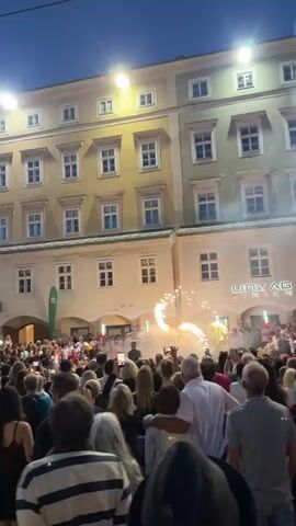 A wide shot captures a crowded street at dusk. A group of people are gathered in the foreground, facing a performance in the background. A building with several floors and numerous windows dominates the background. Below the windows, there is a performance area where a spectacle of fire and sparks is taking place. The crowd appears to be actively watching and recording the event on their phones. The lighting suggests it is evening, with streetlights illuminating the scene and the building's facade. Some smoke is visible from the performance area. The word "LINZ AG" is visible on the building in the background.