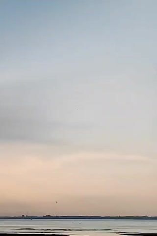 The video shows a tranquil view of a body of water at dusk or dawn. The sky is a blend of pale blue and soft orange, indicative of either a sunrise or sunset.  Clouds are present, with one casting a shadow and rays of light streaming down.  The water surface is calm, reflecting the colors of the sky.  In the foreground, there's a small tree or bush silhouetted against the light. The land surrounding the water appears muddy with various pools and channels.  In the distance, on the horizon, a faint outline of buildings or structures is visible.