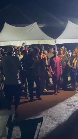 A nighttime outdoor gathering is taking place under white tents. A crowd of people are visible, dressed in various colorful clothing, suggesting a celebratory event. Several individuals appear to be dancing or engaged in lively conversation. In the foreground, a person in a dark shirt and shorts is facing away from the camera, holding a framed object. Another individual in a maroon outfit is also prominent, possibly speaking or being honored. The ground appears wet, indicating recent rain or dampness. The overall atmosphere seems energetic and joyful. The time of day is clearly night, indicated by the darkness and artificial lighting from the tents and possibly some handheld lights. While the specific location within Ufueseni, Nigeria, is not discernible, the setting suggests a community event or festival. No text is visible in the media file.
