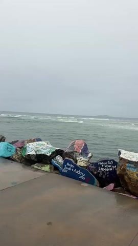 The video shows a section of a rocky shoreline with waves crashing in the background. The rocks are large and unevenly shaped, and many of them are covered in colorful graffiti and writing. The sky is overcast, and the overall mood of the video is somber. There are no people visible in the video. The water appears to be calm, with small waves breaking on the shore. The rocks are arranged in a haphazard manner, creating a natural barrier between the land and the sea. The graffiti on the rocks is varied in style and content, with some of it appearing to be names and dates, while other sections are more abstract designs. The colors used in the graffiti are bright and contrasting, which creates a striking visual effect against the muted tones of the rocks and the sea. The video pans from left to right, showing a panorama of the rocky shoreline.