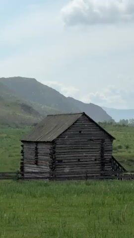 A still, long-shot video displays an old, weathered log cabin in a field of green grass. The cabin has a steeply pitched roof and appears to be made of dark, rough-hewn logs. A wooden fence surrounds the front of the cabin. In the background, rolling hills rise up against a hazy, overcast sky. The lighting is soft and diffused, suggesting it may be a cloudy day. There are no people or animals visible in the frame. The overall impression is one of rustic solitude and historical preservation.