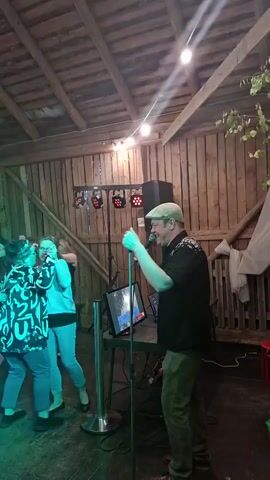 A man in a cap and a patterned shirt sings into a microphone at a karaoke event in a rustic wooden building. He stands to the right of a screen displaying karaoke lyrics. Several people are dancing in the dimly lit space, bathed in colored lights from stage lighting equipment mounted on a truss. The building has exposed wooden beams and walls, with some greenery visible outside on the right.