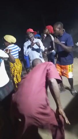 At night, a group of people are dancing outdoors on a dusty surface, possibly at a party. One person is wearing a maroon robe and has their back to the camera. Other individuals are wearing a variety of casual clothing, including a blue and white striped shirt, a white t-shirt, jeans, shorts, and red caps. A woman has a yellow headwrap. The person in the maroon robe is dancing in place, moving their arms and legs. Several people are also dancing. Music is playing in the background. The area appears to be lit by dim artificial light, casting shadows on the ground.