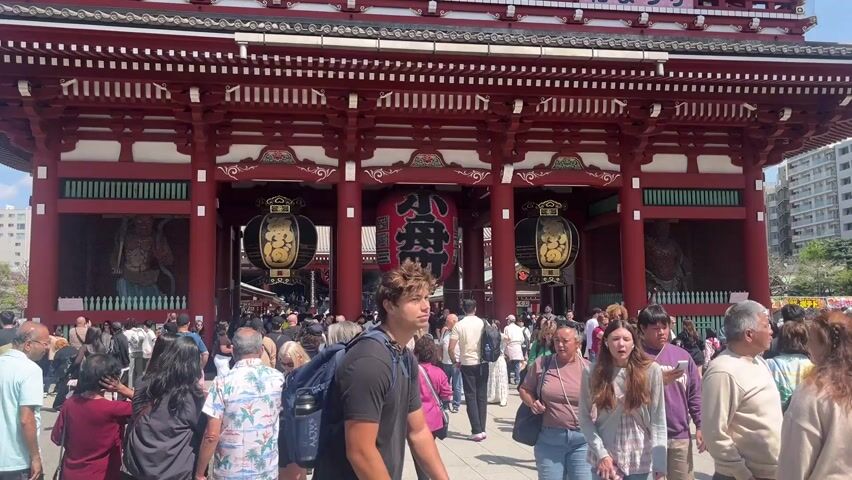 The video shows a large crowd of people in front of a traditional Japanese temple gate. The gate is red and has large lanterns hanging from it. There are statues visible on either side of the gate. People are walking through the gate and milling around in front of it. The sky is blue. Some high rise apartment buildings are visible in the background. A sign with Japanese characters adorns the top of the gate.