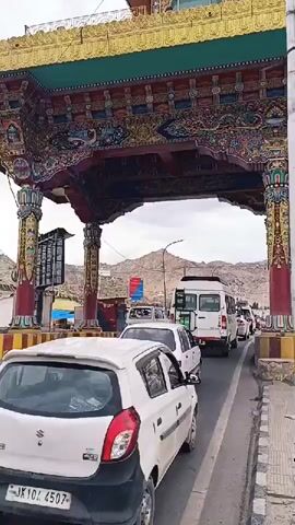 A traffic jam unfolds under a large, ornately decorated archway. A white car is in the foreground, with a bus on its left. Other vehicles, including a white van, are visible further down the road. The archway is painted in vibrant colors, with intricate designs. Mountains are visible in the background. The sky is overcast. The overall impression is one of a busy thoroughfare in a visually striking location.