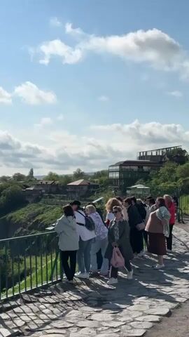 A group of tourists stands behind a green railing at a scenic overlook. The camera pans from the group of tourists, to a stationary telescope, to a dramatic view of a mountain range. The mountains are covered in lush green trees and there are houses built on the cliffs. The sky is blue and partly cloudy.