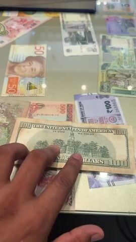 A close-up shot of a hand holding a US 100 dollar bill over other currencies. The hand is holding the bill at the top edge, with the face of Benjamin Franklin visible. The 100 dollar bill is placed on top of several other banknotes, including what appears to be Indian rupees and possibly other international currencies. The background shows more banknotes spread out on a reflective surface, possibly a glass table. The lighting suggests an indoor environment, possibly a shop or a display area for currencies. There is also a hint of a calculator or electronic device in the upper right corner and a book or ledger in the upper left corner, suggesting a commercial or transactional setting. The focus is primarily on the 100 dollar bill and the hand holding it.