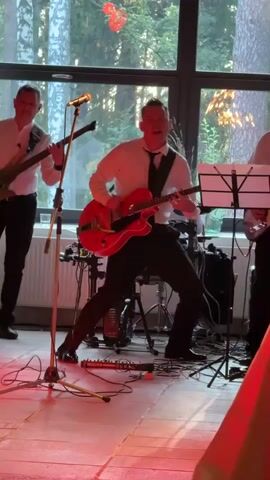 A band is playing music indoors. Three men are visible, two are playing guitars and one is playing drums. The man in the center is playing a red electric guitar and singing into a microphone. He is wearing a white shirt, black tie, and black pants. The man on the left is playing a bass guitar and is wearing a white shirt and black pants. The man on the right is playing a guitar and is wearing a white shirt and black pants. There are drum set behind the guitarists. The room has large windows that look out onto trees and sunlight. Red and white lights are illuminating the stage floor.