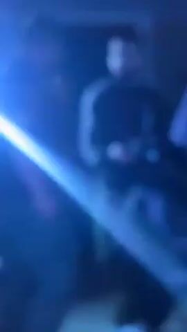 Two men are looking at the screen of a camera in a room illuminated by blue light. The man on the right is wearing a black tracksuit with three white stripes and has a beard. He is holding the camera. The man on the left is wearing glasses and a black t-shirt. A blue cloth hangs off to the right.