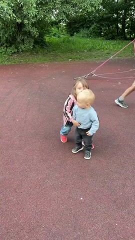 A young girl stands behind a baby on a playground. The girl is wearing a pink and black jacket, blue jeans, and red shoes. The baby is wearing a light blue long-sleeved shirt, black pants, and black shoes. The girl has her arms around the baby and they are both facing forward. The playground has a red rubber surface and a grassy area with trees in the background. The girl briefly turns her head to the left and then points to her right. The baby then walks to the right.