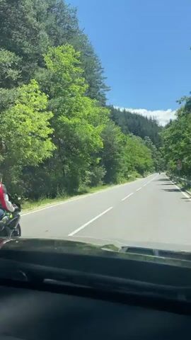 The video shows a view from inside a car looking out at a paved road winding through a lush green forest on a sunny day. A person on a motorcycle, wearing a red helmet and a red backpack, is riding ahead on the road. The road is flanked by dense trees and foliage, and there is a clear blue sky above. The car appears to be following the motorcycle at a distance, and another motorcycle is visible further ahead in the distance. The car camera pans slightly, offering a view of the road, the surrounding forest, and other vehicles. A speed limit sign indicating 50 is visible on the right side of the road.