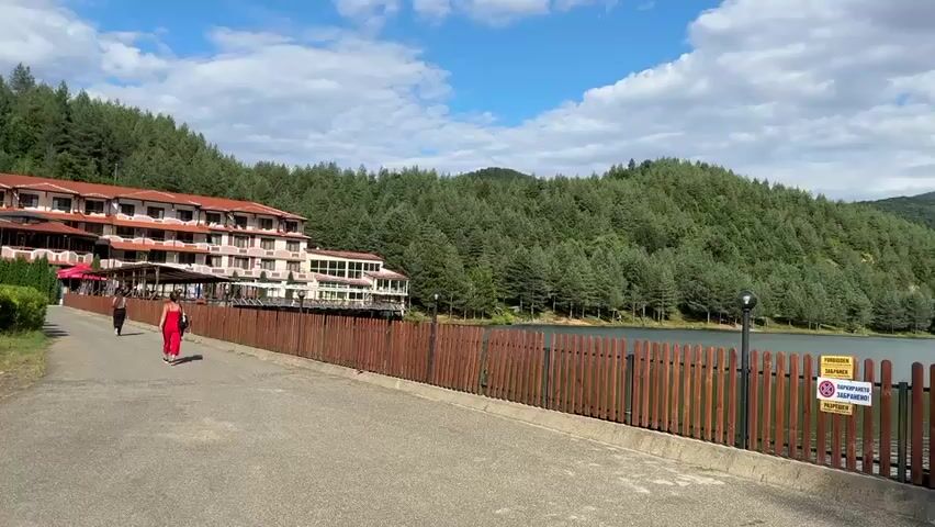 The video pans across a scenic view of a hotel nestled beside a lake and surrounded by lush green forested hills under a partly cloudy sky. In the foreground, a paved path runs alongside a wooden fence. Two individuals are walking on the path, one in a red dress and another in dark clothing. A parked car is visible on the left side of the path. The lake is calm, reflecting the sky and the trees. Several signs are posted on the fence, and a security camera is mounted on a pole near a small building on the right side of the frame. The overall atmosphere is serene and picturesque.