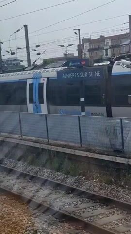 The video shows a train at a station. The train is blue and white. There is a fence between the train and the tracks. There are buildings in the background. The video is shaky, suggesting it was filmed from a moving vehicle.