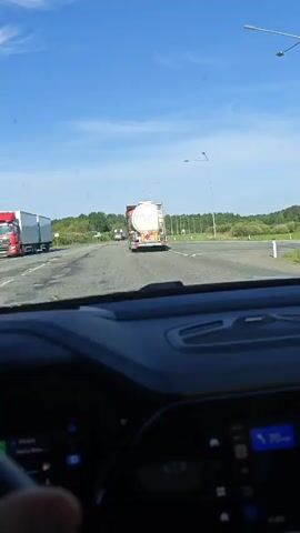 A driver is following two trucks on a divided highway. The first truck appears to be a tanker, and the second is obscured by the tanker. The weather is clear and sunny. The dashboard of the car is visible. The car is traveling at 70 km/h. Trees and vegetation line the side of the road. A street light is visible.