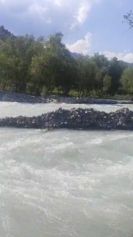 A video shows a flowing river with a rocky shore on the left and trees and mountains in the background. The water is light-colored and appears to be moving quickly, with some ripples and whitewater visible. Several large rocks and stones are present in the riverbed and along the shore. The sky is partly cloudy, with sunlight illuminating the scene. The video pans slightly across the river and the surrounding landscape.
