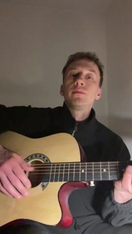 A fair-skinned man with short brown hair is sitting and playing an acoustic guitar. He is wearing a black zip-up jacket. The guitar has a natural wood finish, a black pickguard, and a red burst color on the body where it meets the neck. The background is a plain light grey wall. The man is looking directly at the camera as he plays the guitar.