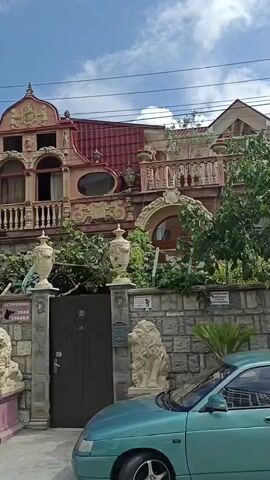 This is a video of a large, ornate house with a red tiled roof. The house has many decorative elements, including columns, balconies, and a central tower-like structure. In front of the house is a stone wall with two lion statues guarding a dark colored gate. A turquoise car is parked in front of the wall and gate. The sky is blue with some clouds. The overall impression is that of a very grand and possibly eccentric property.
