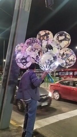 The image depicts an urban night scene, likely in Maipú, Chile. A man, seen from his right side and back, stands on a sidewalk holding a large cluster of approximately ten to twelve clear, bubble-like balloons. He is dressed in a dark jacket or shirt over blue jeans.

The balloons are the central focus, glowing brightly from internal LED lights that emit a vibrant mix of purple, pink, and white hues. Each transparent balloon contains various internal decorations, such as confetti, smaller balloons, or distinct shapes and figures. The balloons are held together by a bunch of green and blue sticks or handles that the man grasps with his right hand.

The setting is a street at night, indicated by the darkness and artificial lighting. A silver or white car is partially visible on the right side of the frame, appearing to move past the man on the road. In the blurred background, other vehicle lights and urban structures are faintly discernible, suggesting a busy thoroughfare. The man's posture, holding the prominent, illuminated balloons, implies he might be a street vendor displaying his merchandise. There is faint, mostly unreadable text on some of the balloons.