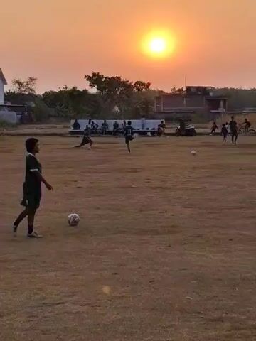 This video captures a group of people playing soccer on a dusty field during sunset. The sun, a bright orange orb, is prominently positioned in the sky, casting a warm glow over the scene. Several individuals are actively engaged in the game, including one person in the foreground wearing a green jersey and shorts, who is positioned with a soccer ball at their feet. Other players are scattered across the field, some running, kicking the ball, or in defensive stances. In the background, a line of spectators sits along the edge of the field, observing the game. Beyond the field, a few buildings and trees are visible, silhouetted against the vibrant sunset sky. The overall atmosphere appears to be one of casual recreation and community engagement.